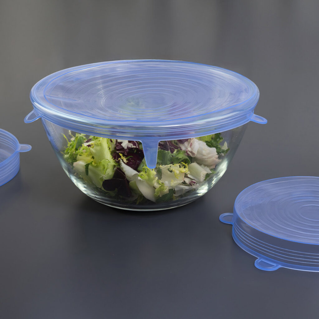 Eine mit Salat gefüllte Glasschale ist mit einem blauen, wiederverwendbaren Silikon-Dehndeckel abgedeckt. Ein weiterer Silikondeckel liegt flach daneben auf einer dunklen Fläche.