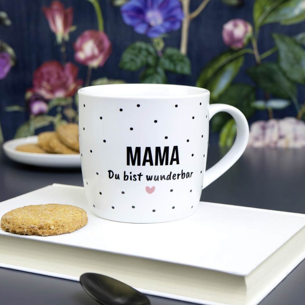 Ein weißer Becher mit schwarzen Punkten und dem Schriftzug MAMA Du bist wunderbar steht auf einem geschlossenen Buch neben einem Keks, mit weiteren Keksen und einer Blumentapete im Hintergrund.