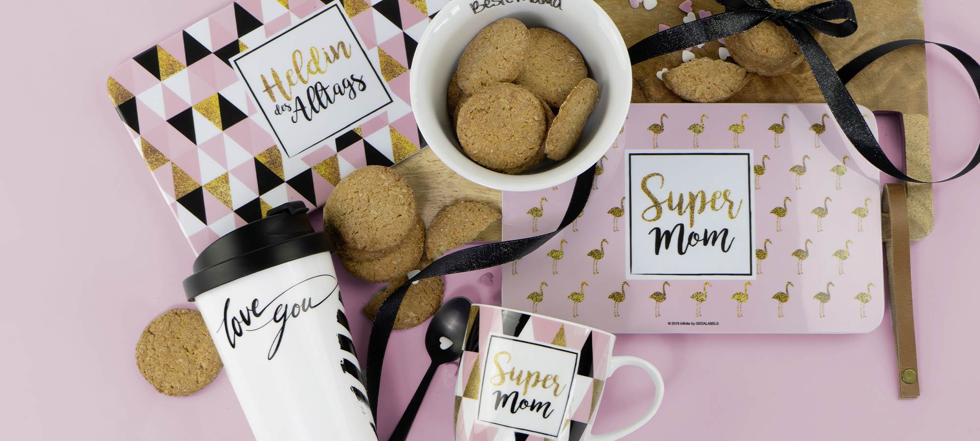 Geschenkset auf rosafarbenem Hintergrund mit Keksen, einer Super Mom Tasse und einem Notizbuch, einem Holdin it all together Klemmbrett, einer Love you Reisetasse, einer Best Mom Schale und verstreuten Keksen mit schwarzem Band.