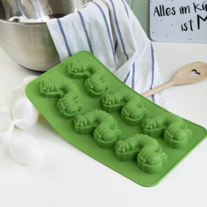Eine grüne Silikonform mit acht Raupenformen liegt auf einer Arbeitsplatte neben weißen Eiern, einem Holzlöffel mit Herzausschnitt, einem gestreiften Handtuch und einer Metallschüssel.