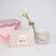Eine rosafarbene Geschenkbox, ein rosafarbener Umschlag, eine weiße Tasse mit der Aufschrift "Heldin des Alltags", eine Vase mit weißen Blumen und eine Karte mit der Aufschrift "Super Mom", umgeben von kleinen Flamingos.